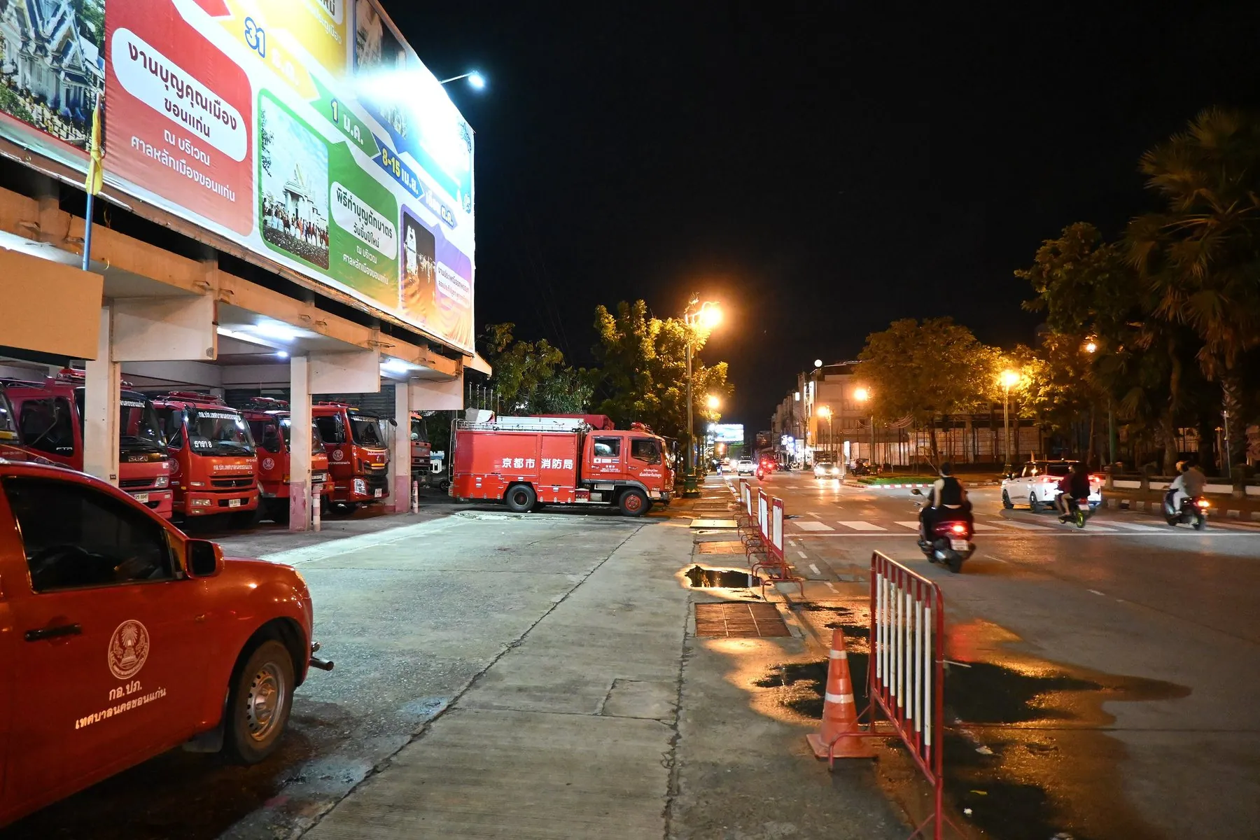 Red fire engines and a rescue-foundation pickup standing in the open bays of a Thai fire station at night as motorbikes pass on the wet road