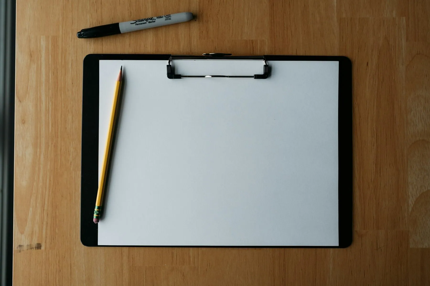 A clipboard holding a blank sheet, with a pencil and a marker pen lying beside it on a wooden surface