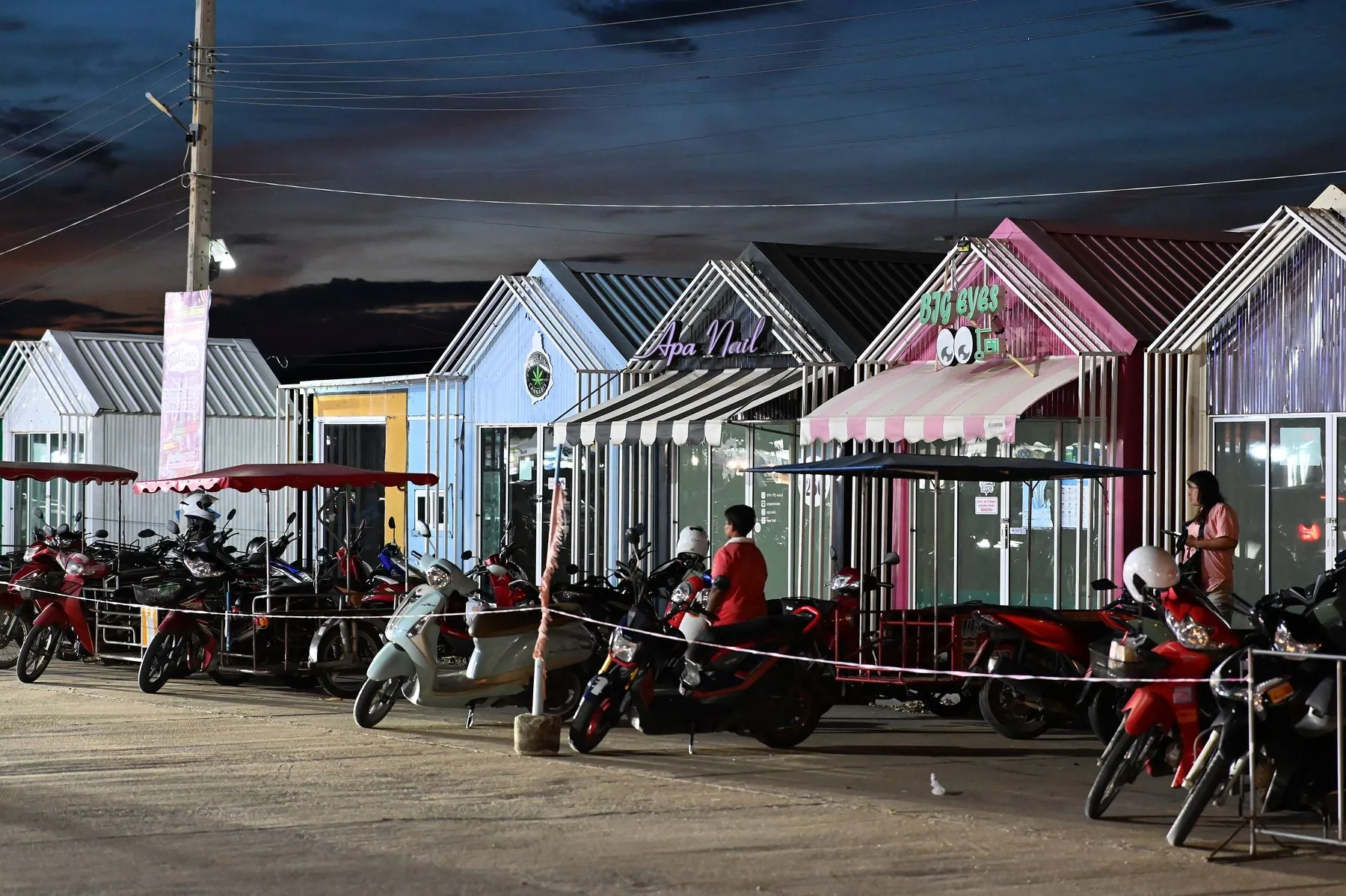 Scooters and motorbikes parked nose-in along a rope barrier in front of a row of small Thai beach shops
