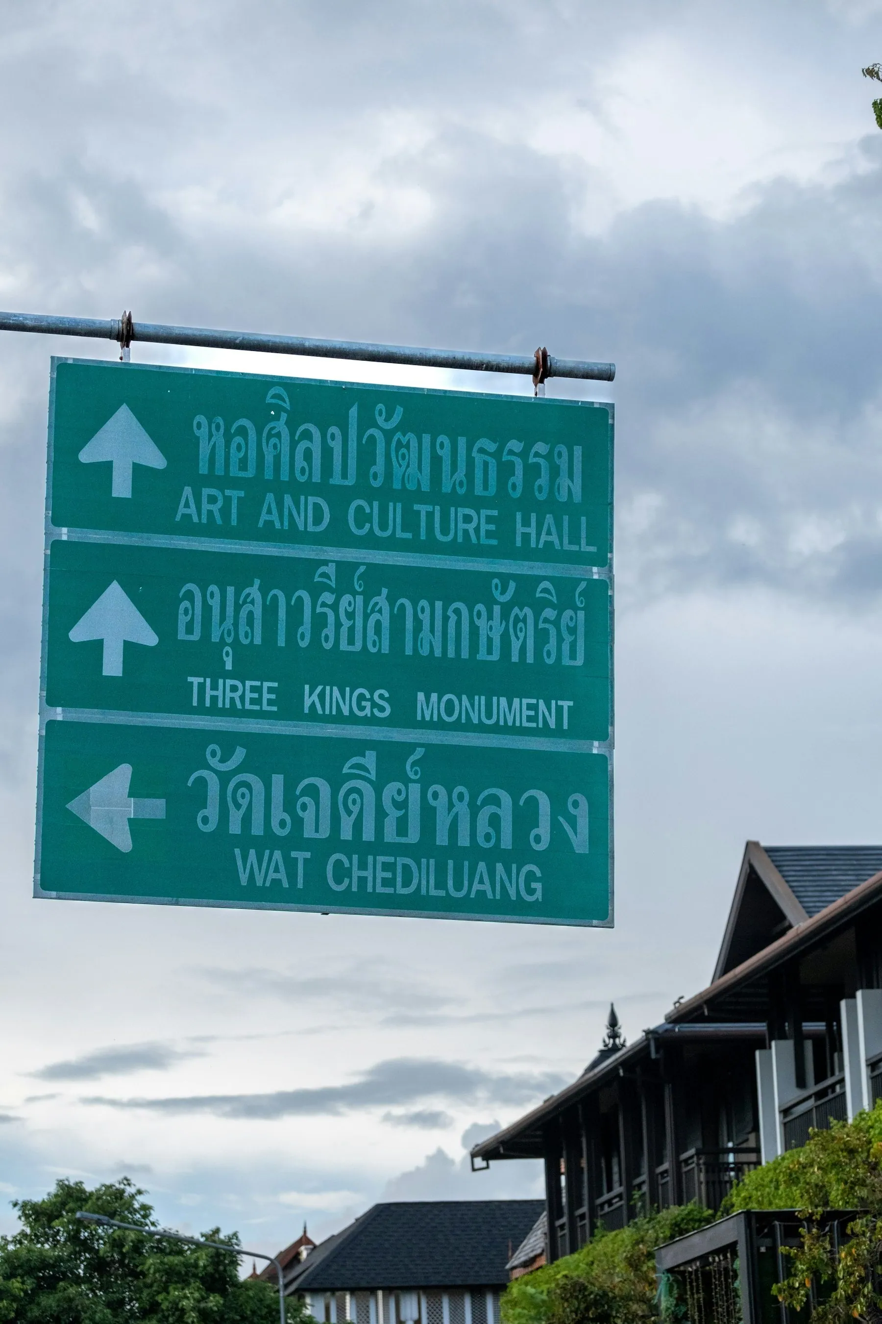 Stacked green direction signs in Thai and English pointing to the Three Kings Monument and Wat Chedi Luang in Chiang Mai