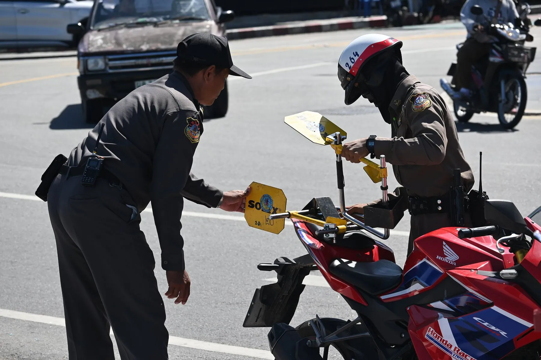 Two Thai traffic officers bending over a red motorcycle at the kerb to fit a yellow wheel lock to its front wheel