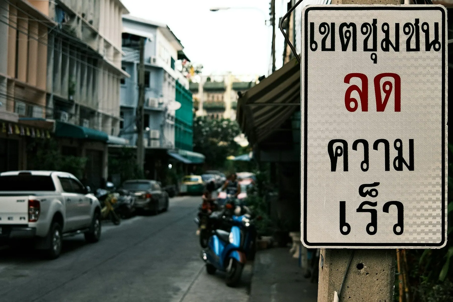 A white Thai-script sign warning drivers to slow through a built-up area, above a street of pickups and parked scooters