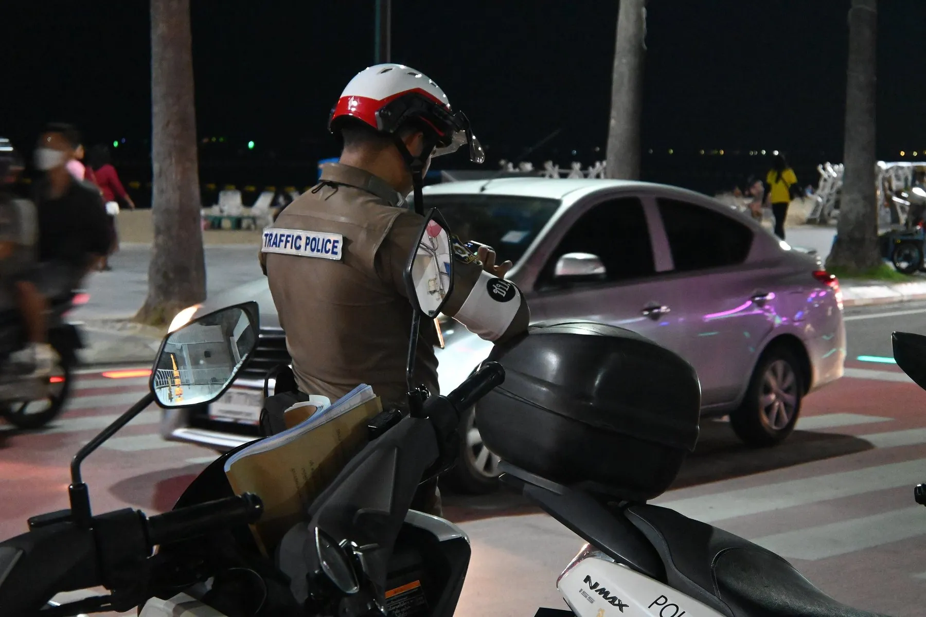 A Thai traffic police officer in a white helmet reads a driver's papers from the saddle of his police motorbike beside a stopped car at night